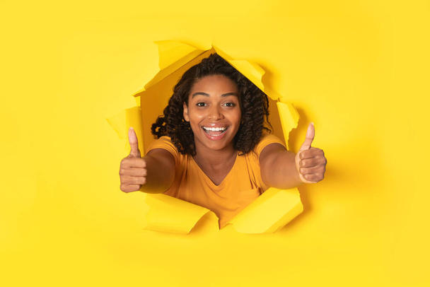 Excited Black Lady Gesturing Thumbs Up Stretching Arms To Camera Through Hole In Torn Paper Approving Great Offer Posing On Yellow Background In Studio. I Like It Concept - Photo, Image