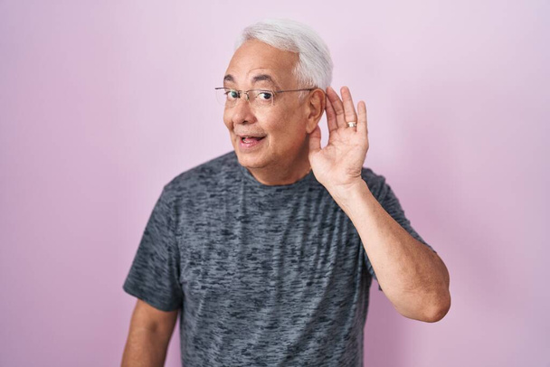噂やゴシップに耳を傾ける耳の上に手で笑みを浮かべてピンクの背景に立って灰色の髪を持つ中年の男。聴覚障害の概念.  - 写真・画像