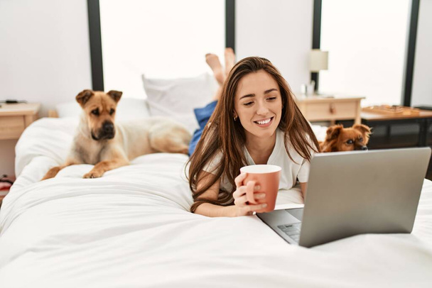 Young hispanic woman using laptop and drinking coffee lying on bed with dogs at bedroom - Photo, Image