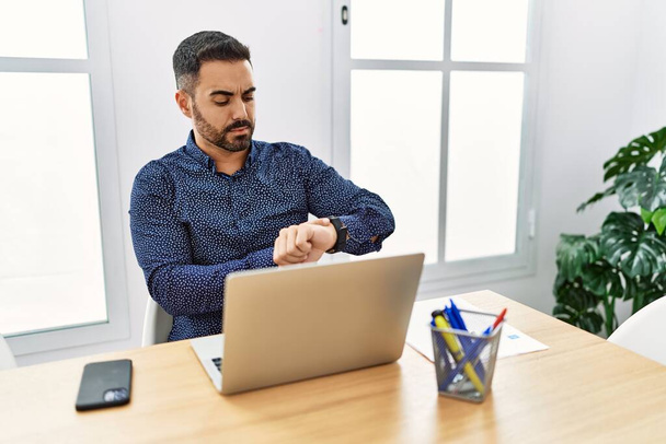 Young hispanic man with beard working at the office with laptop checking the time on wrist watch, relaxed and confident  - Foto, imagen
