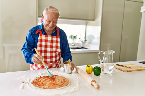 Hombre mayor sonriendo confiado cocinar pizza en la cocina - Foto, Imagen
