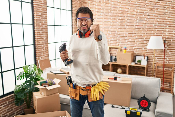 Handsome middle age man holding screwdriver at new home angry and mad raising fist frustrated and furious while shouting with anger. rage and aggressive concept.  - Photo, Image