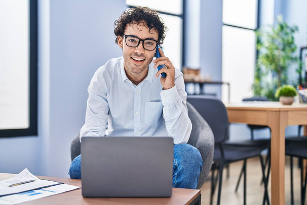 Young hispanic man business worker using laptop talking on smartphone at office - Valokuva, kuva