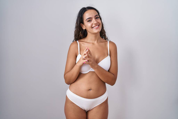 Молодая испаноязычная женщина в белых руках нижнего белья вместе, а пальцы скрещены, улыбаясь расслабленной и веселой. успех и оптимизм  - Фото, изображение