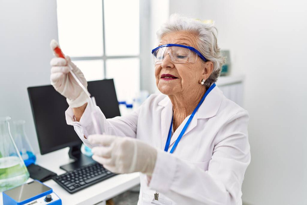 Oudere grijsharige vrouw in uniform met pipet en reageerbuis in het laboratorium - Foto, afbeelding