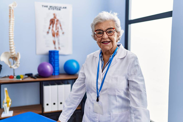 Mujer mayor de cabello gris que usa uniforme de fisioterapeuta de pie en la clínica de fisioterapia - Foto, Imagen