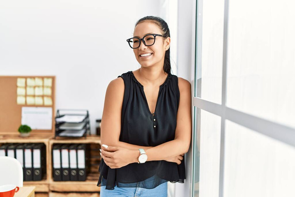 Young latin woman smiling confident standing with arms crossed gesture at office - 写真・画像
