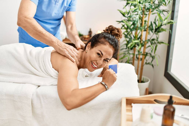 Middle age man and woman wearing therapist uniform having back massage session and holding credit card at beauty center - Foto, Imagen
