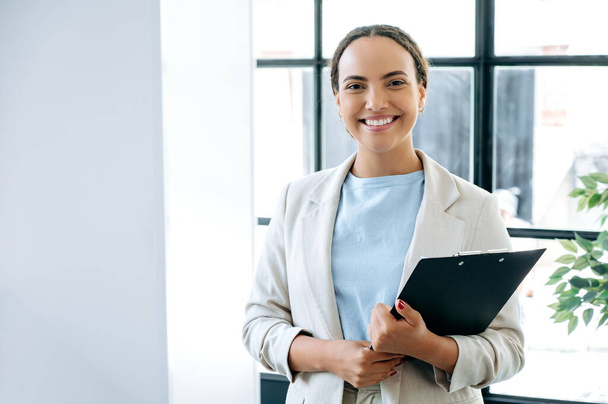 Photo of a successful confident positive brazilian or latin business woman standing in a working office with a folder of documents in her hands, financial manager, realtor, looking at camera, smiles - Foto, afbeelding