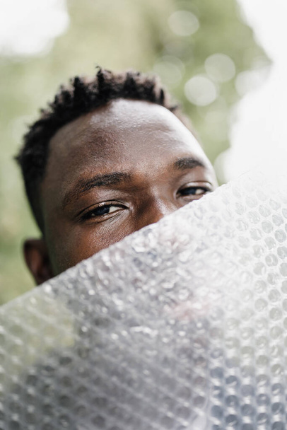 Young African American man outdoor, close-up street portrait with plastic bubble wrap foil. Ecology, environmental conversation, mental health concept. Fall or spring season - Photo, Image
