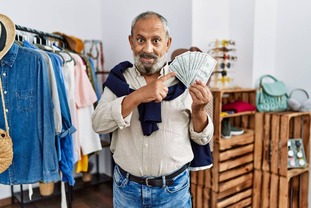 Handsome senior man at retail shop holding dollars banknotes smiling happy pointing with hand and finger  - Photo, Image