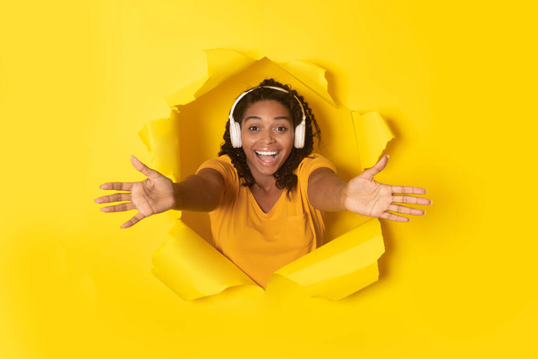 Радостная чёрная женщина носит наушники, растягивающие руки к камере, слушая музыку, позирующую в дыре в порванной бумаге на жёлтом фоне. Великий плейлист и музыкальные гаджеты - Фото, изображение