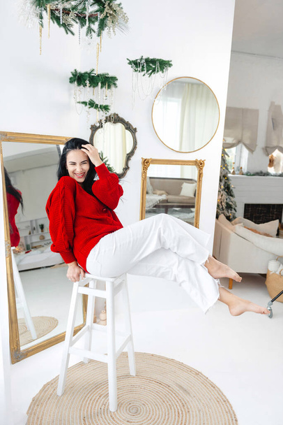 A beautiful girl in light-colored pants and a red sweater stands in front of a Christmas atmosphere. The girl is laughing and in a good New Years mood - Φωτογραφία, εικόνα