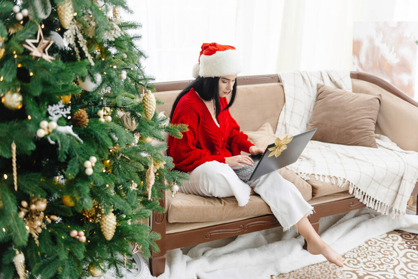 The girl with the laptop. A beautiful woman wearing a red Christmas hat on her head does online shopping on the Internet through her laptop. Christmas discounts. Christmas atmosphere and Christmas - Foto, immagini