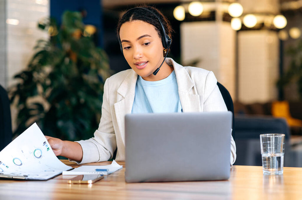 Busy focused brazilian or hispanic businesswoman, consultant, call center worker, with headset, sitting at work desk in office, remotely consulting client, giving recommendations, answering questions - Photo, Image