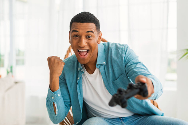 Satisfied excited young african american male with joystick playing online game, rejoices to win, sits on chair in lite living room interior. Gesture of success and victory, entertainment at home - Photo, Image