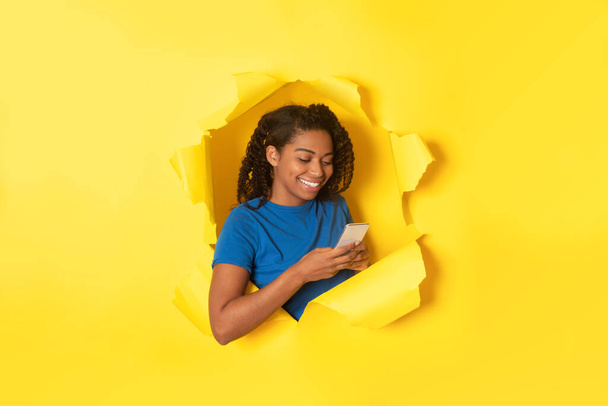 Sorrindo mulher afro-americana usando aplicação em mensagens de texto de celular posando no buraco em papel amarelo rasgado, Studio Shot. Comunicação móvel e conceito de tecnologia. Colagem - Foto, Imagem