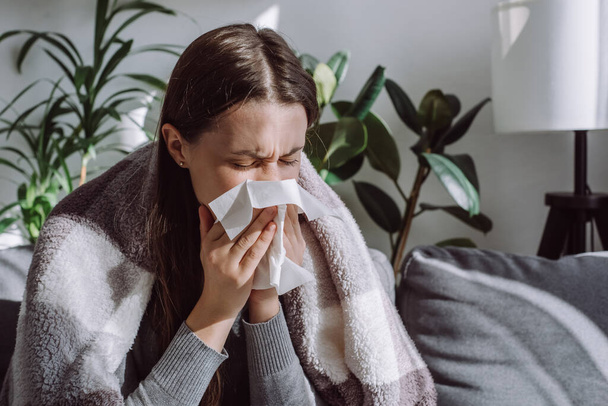 Close up of sick woman sitting on sofa freezing blowing running nose got fever caught cold sneezing in tissue, ill brunette girl covered with blanket, having influenza symptoms coughing at home - Фото, изображение
