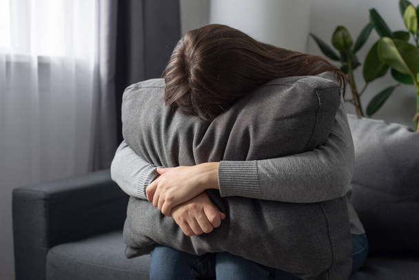 Depressed single young woman holding pillow going through emotional crisis after abuse abortion sitting on couch at alone home. Lonely stressed female in sofa hide from problems of hard cruel world - 写真・画像
