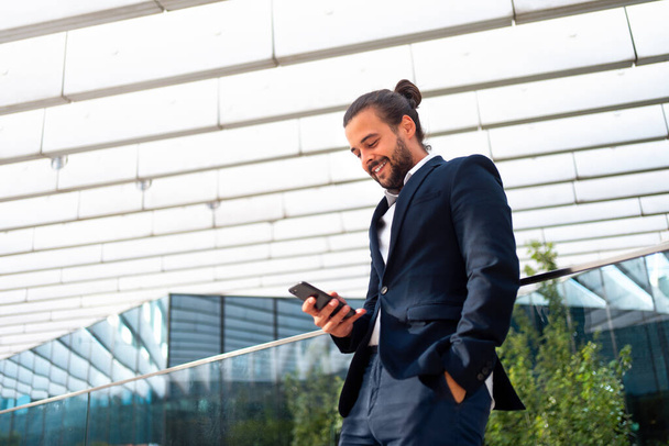 Businessman standing outdoor use smartphone looking device screen and smile. Handsome hispanic male business person in business suit holding smartphone in hand. Modern business person low angle view - Фото, зображення