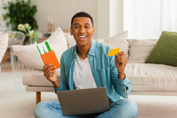 Gai millénial homme afro-américain avec ordinateur portable montre passeport, billet d'avion et carte de crédit, se réjouit dans le voyage et le cashback dans l'intérieur du salon. Vacances, voyages, tourisme et paiement à distance - Photo, image