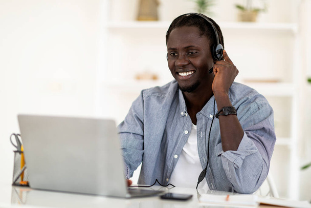 Travail à distance. Heureux mâle noir portant casque en utilisant un ordinateur portable pour le travail en ligne, sourire jeune homme afro-américain dans les écouteurs assis au bureau à la maison et de travailler sur ordinateur, Copier l'espace - Photo, image