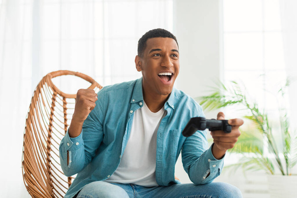 Gusto emocionado millennial afroamericano chico con joystick jugando juego en línea, se regocija en la victoria, se sienta en la silla en el interior de la sala de estar blanca. Gamer disfruta de dispositivo, hace gesto de éxito y ganar - Foto, imagen