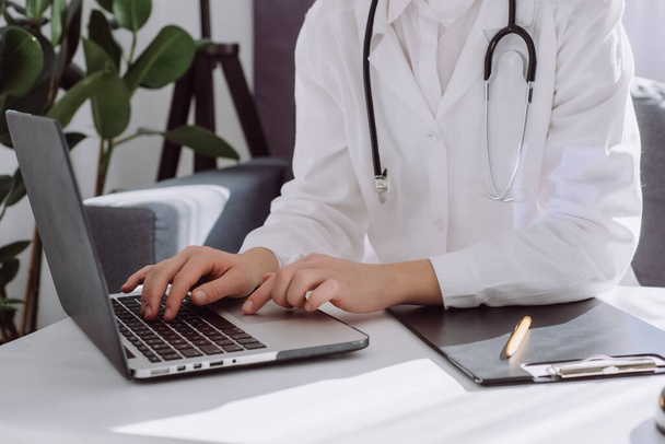 Close up of professional young woman doctor in white coat with stethoscope using laptop sitting on comfy couch at desk in clinic, nurse working on computer at room office. Medical work concept - Φωτογραφία, εικόνα