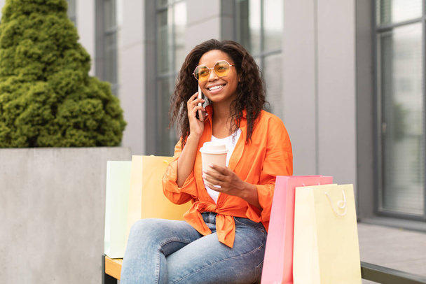 Fröhliche junge afrikanisch-amerikanische Frau in Lässigkeit und Brille mit vielen Taschen ruft per Smartphone auf der Straße der Stadt an. Tolles Angebot und Werbung, Shopaholic genießt Shopping, Klatsch, gute Nachrichten und Kommunikation - Foto, Bild
