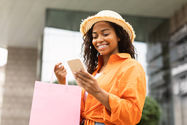 Szczęśliwa młoda, ładna, afrykańska kobieta w luźnym kapeluszu z wieloma torbami z zakupami na smartfonie, używająca bankowości na ulicy miejskiej. Nowa aplikacja zakupy online, zakupoholiczka cieszyć sprzedaż i blog mody - Zdjęcie, obraz