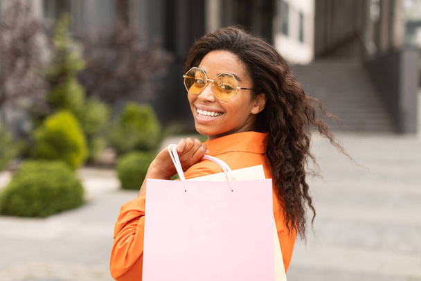 Portrait de jeune femme afro-américaine souriante en casual et lunettes avec sac avec achats, regardant la caméra dans la rue de la ville, gros plan. Grande offre et annonce, shopaholic profiter du shopping et de la vente - Photo, image