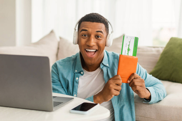 Glad excited young african american guy in wireless headphones with passport, air tickets, near computer, enjoys order in living room interior. Vacation, trip, journey after covid-19, people emotions - Photo, Image