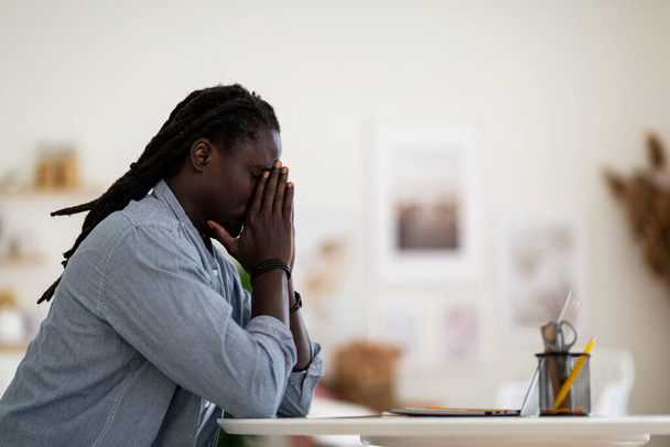 Πλευρική άποψη του θλιμμένος νεαρός Αφροαμερικανός που κάθεται στο γραφείο με το φορητό υπολογιστή στο σπίτι, αναστατωμένος μαύρος τύπος που πάσχουν από προβλήματα ζωής ή άγχος στην εργασία, κρατώντας σφιγμένα χέρια κοντά στο πρόσωπο, αντιγραφή χώρου - Φωτογραφία, εικόνα