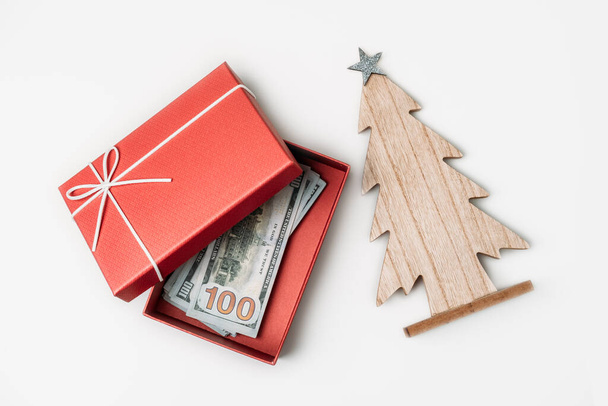 Πώς να περάσετε, τον προϋπολογισμό των Χριστουγέννων και την εξοικονόμηση έννοια. Χριστουγεννιάτικο κουτί δώρου, στοίβα από χαρτονομίσματα δολαρίων και ξύλινο χριστουγεννιάτικο δέντρο σε λευκό τραπέζι. - Φωτογραφία, εικόνα