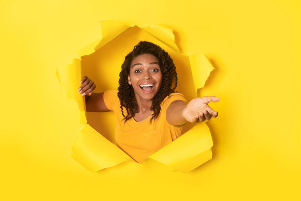 Give Me Your Hand. Excited African American Lady Stretching Hand To Camera Through Hole In Torn Paper Posing On Yellow Background In Studio. Great Offer Advertisement Concept. Collage - Photo, Image