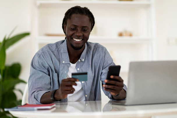 電子商取引の概念。笑顔の黒人男性クレジットカードを保持し、自宅のオフィスで机に座っている間に携帯電話を使用して、幸せなアフリカ系アメリカ人男性フリーランサーオンライン決済を行う、スペースをコピー - 写真・画像