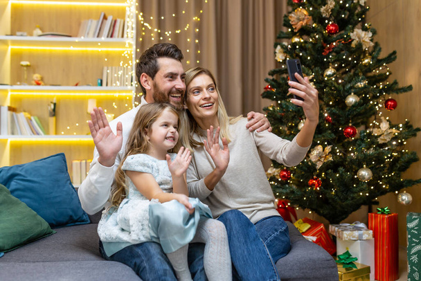 Happy family celebrating new year and christmas, husband wife and little daughter sitting on sofa in living room and talking with relatives on video call, using phone for remote communication. - Valokuva, kuva