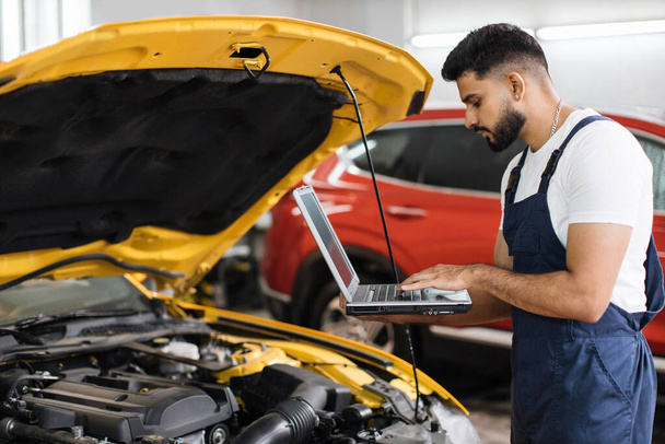 Bearded male mechanic using laptop, recording automobile engine checks collect detailed information during his work on car workshop. Service maintenance during engine repair. - Foto, Imagem