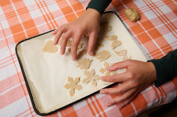Von oben betrachtet Hände einer Hausfrau, Konditorin, die geschnittene Formen aus rohem Teig auf das Backblech legt, bevor sie Lebkuchen in den heißen Ofen legt. Weihnachtliches Kulinarisches und Gebäck - Foto, Bild