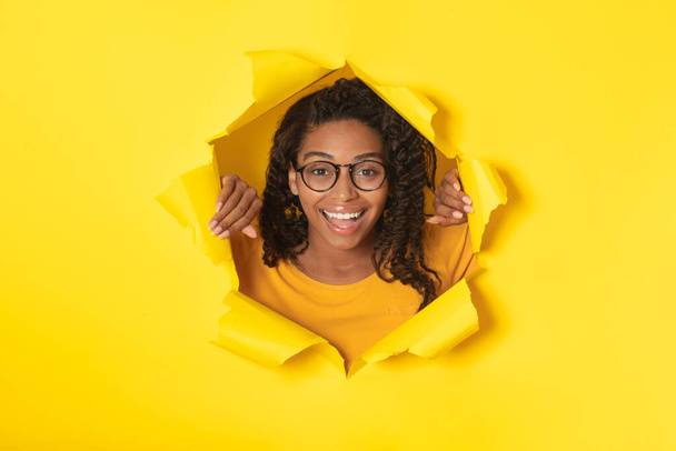 Yırtık kağıda poz vermiş, sarı stüdyodaki delikten kameraya gülümseyen mutlu siyah kadın. Büyük Teklif Reklam Konsepti. Kolaj - Fotoğraf, Görsel