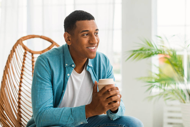 Smiling millennial african american guy with cup of coffee takeaway, sit on chair, enjoy comfort and favorite drink in white living room interior. Rest and relaxation, coffee break at home, covid-19 - Photo, Image