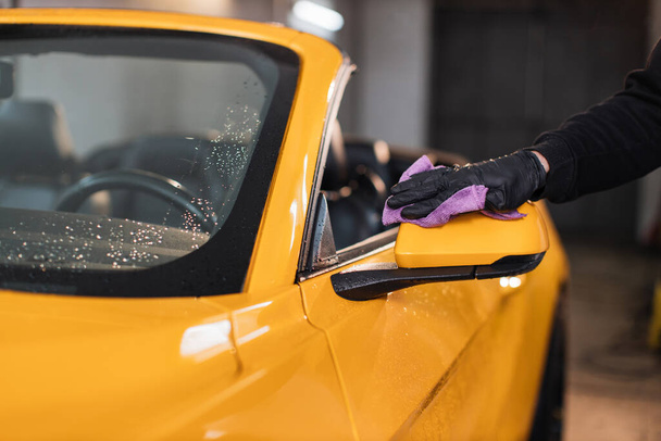 Close up of hand of male car service worker hand in black rubber glove holding the microfiber cloth and cleaning car mirror at modern car wash service. - Фото, изображение