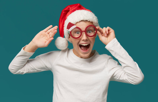Cute teen boy in a Santa Claus hat and party glasses on a turquoise background. The child smiles in surprise with his mouth wide open. wow funny face - Zdjęcie, obraz