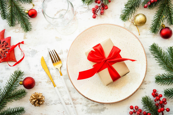 Świąteczne jedzenie, nakrycie stołu Bożego Narodzenia z białym talerzem, złote sztućce, pudełko na prezenty i dekoracje świąteczne na białym drewnianym tle. Widok z góry. - Zdjęcie, obraz