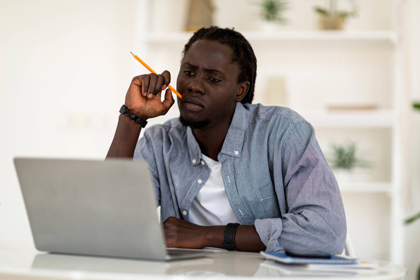 Przemyślany młody Afroamerykanin trzymający ołówek i patrzący na ekran laptopa, zamyślony czarny mężczyzna freelancer siedzący przy biurku z komputerem, pracujący nad nowym projektem w biurze wewnętrznym, wolna przestrzeń - Zdjęcie, obraz