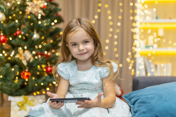Portrait of a little girl at home with a smartphone sitting on the sofa near the Christmas tree watching video content online on Christmas and New Years holidays, looking at the camera and smiling. - Фото, изображение