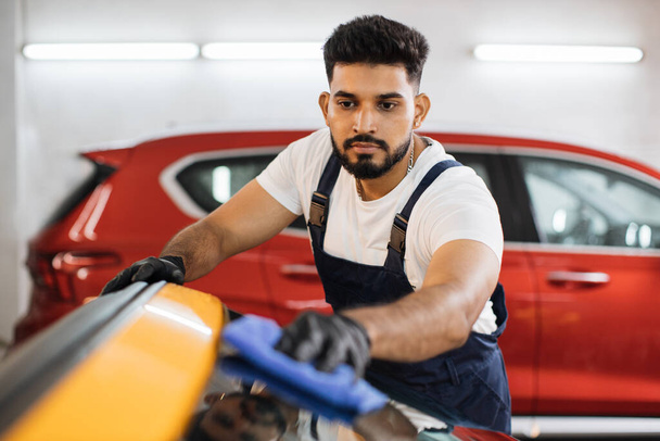 Bearded car wash worker man, wearing protective gloves, cleaning luxury modern yellow car window with microfiber cloth. Car detailing or vehicle concept. - Fotoğraf, Görsel
