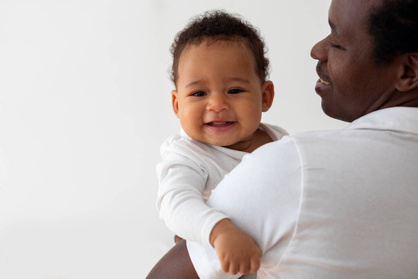 Junger glücklicher Vater, der liebenswert lächelndes kleines schwarzes Baby in den Armen hält, Millennial-Afroamerikaner, der sich mit seinem niedlichen Kleinkind zu Hause verbindet, fröhlicher Junge, der in die Kamera blickt, Nahaufnahme - Foto, Bild