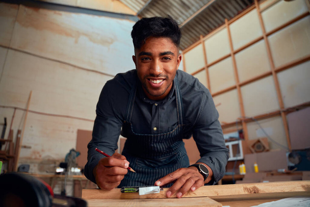 Portrait of multiracial smiling young man holding pencil and using tool on wood at woodworking factory - Фото, зображення