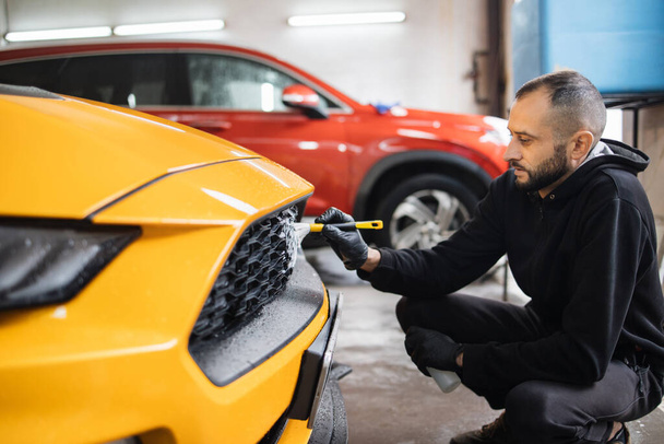 Car detailing wash. Manual cleaning with soap at car wash. Portrait of bearded male worker in rubber glove washing car radiator grille of luxury yellow car with special brush and cleansing foam. - Fotó, kép